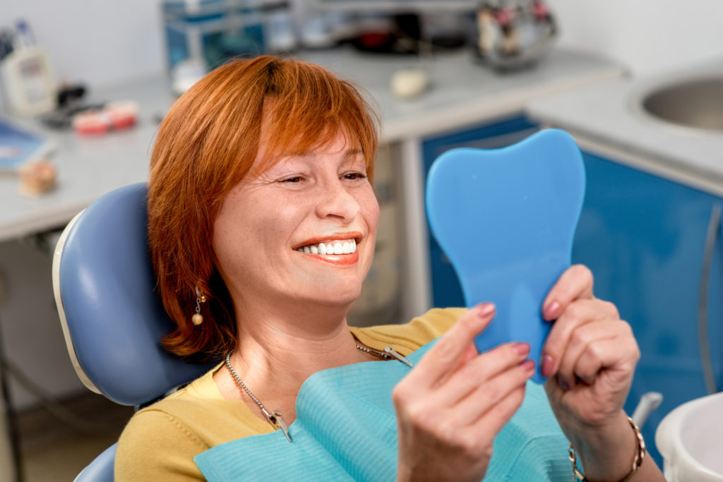 A woman smiling in a mirror sitting in a dental chair happy with her results.