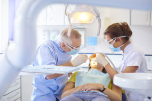 A senior dentist scrapes away some debris from between a patient tooth while the dental nurse applies suction in the mouth to take away any foreign objects. The brightly lit dentist surgery is clean and modern.